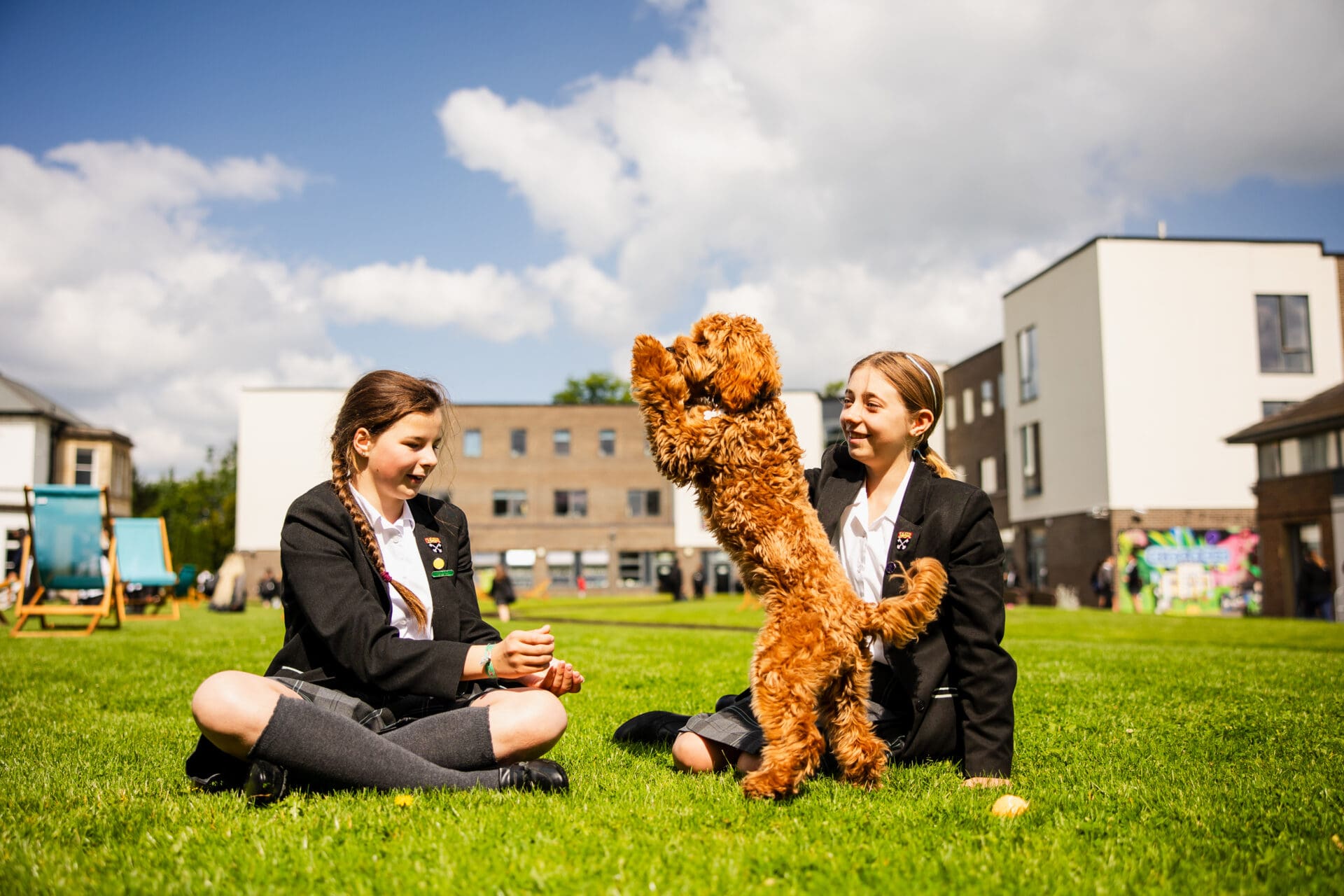 Image of 2 female pupils sat on the grass with one of the school dogs