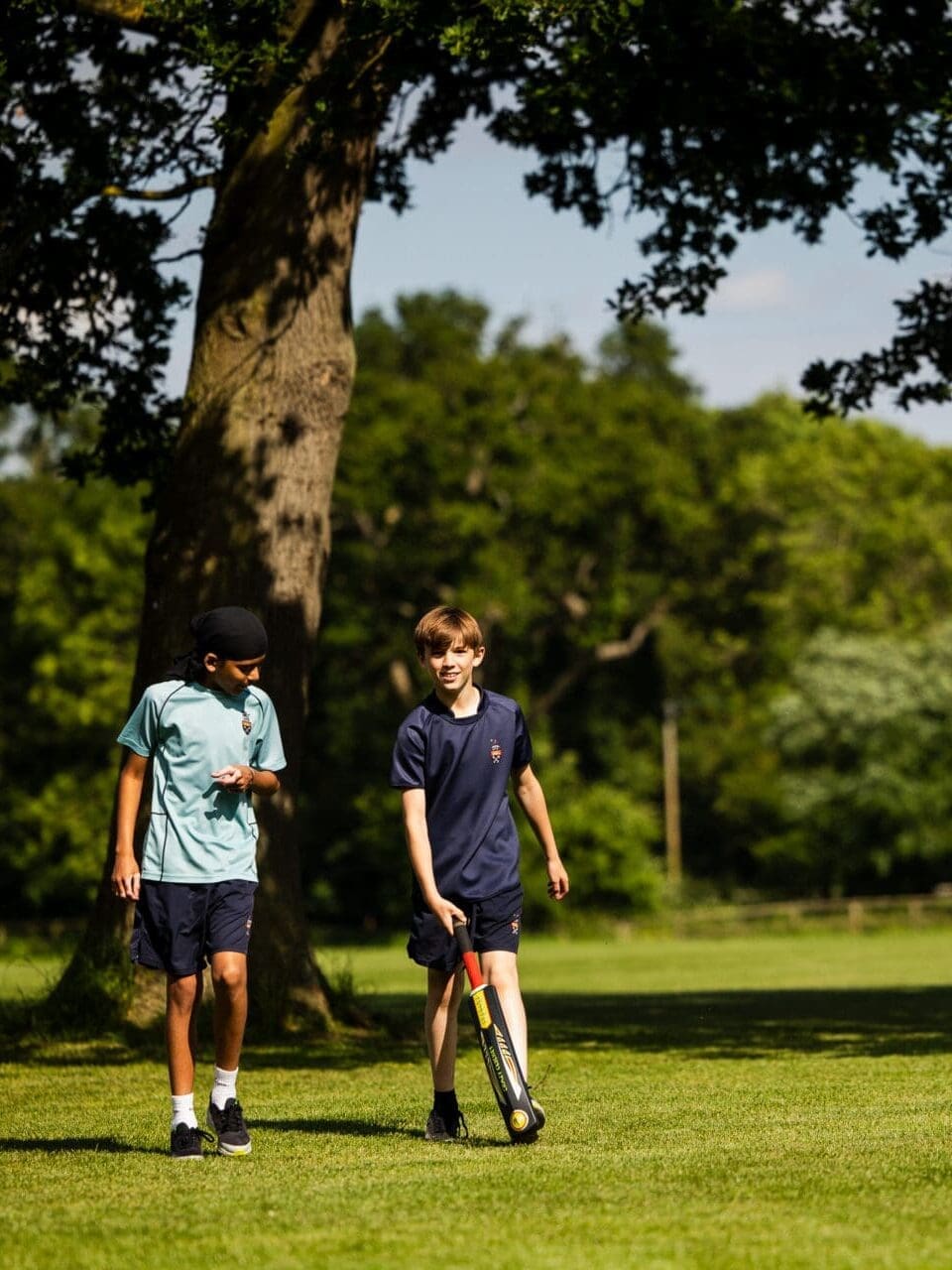 Image of 2 male students walking through the playing fields with a cricket bat